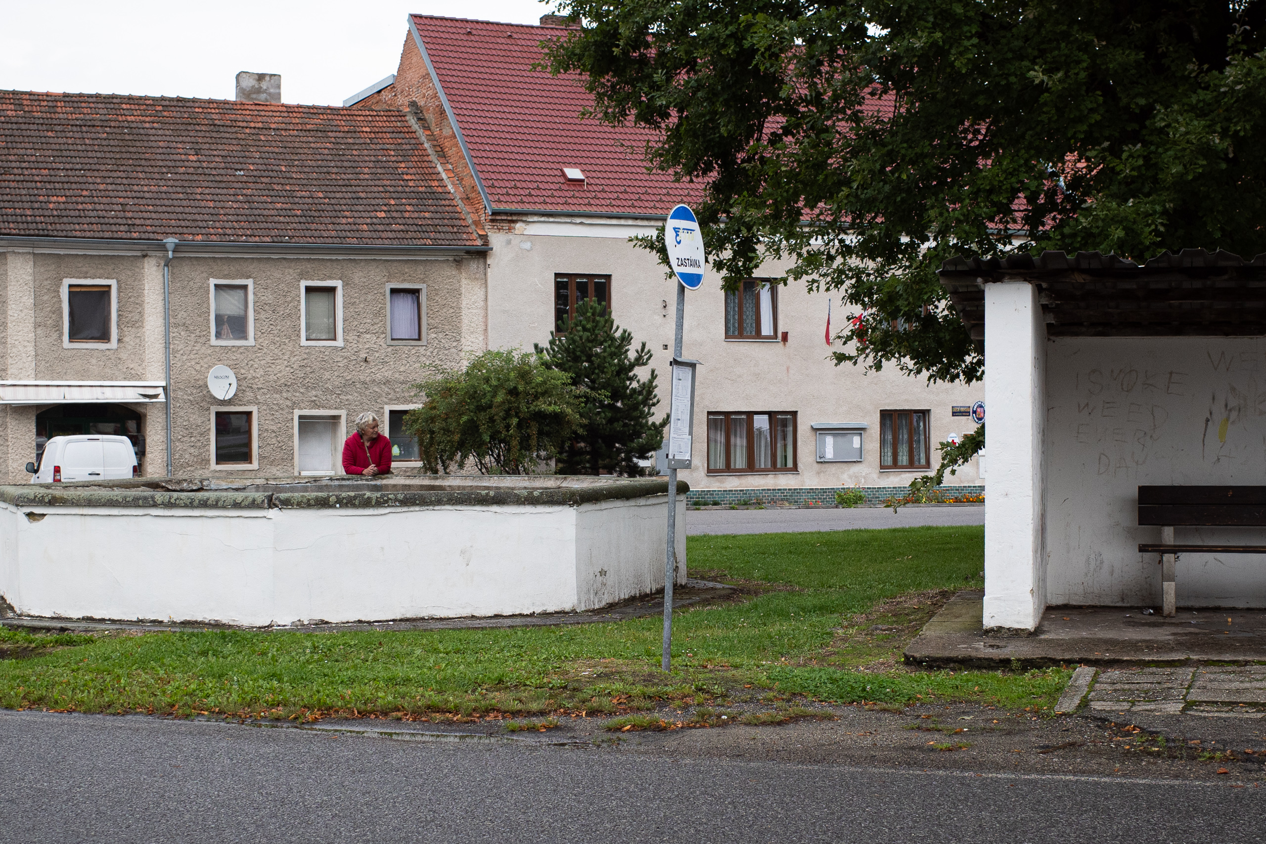 Dorfplatz mit Bushaltestelle – eine Frau lehnt am Rand des Beckens
